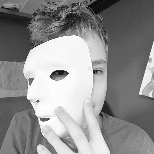 Black and white photo of researchers son holding a mask to his face while looking through one eye of the mask at the camera.
