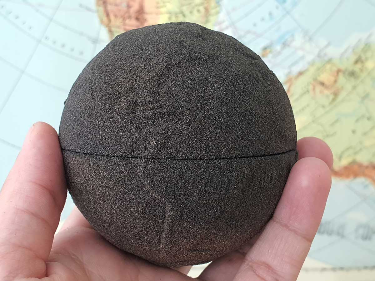A person’s hand holding a dark brown, spherical object with a seam around the middle against a blurred background featuring part of a world map.
