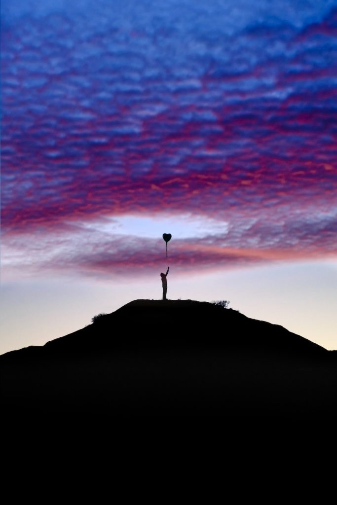 A silhouetted figure atop a hill reaches for a heart-shaped balloon against a purple and blue twilight sky.