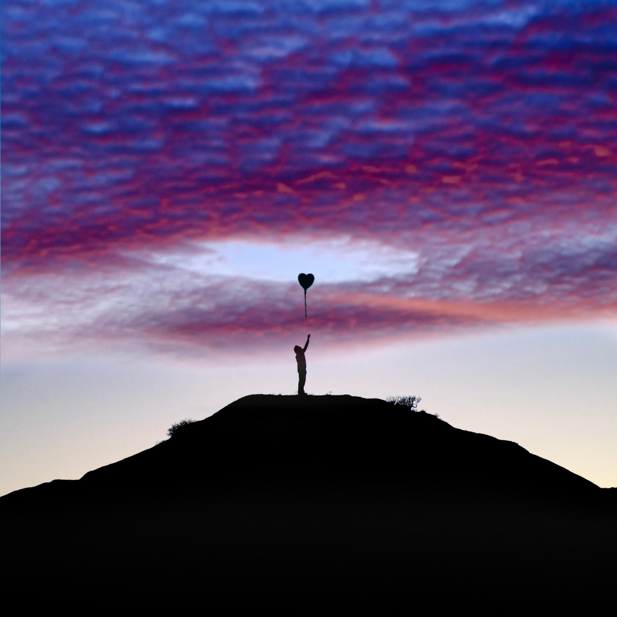 A silhouetted figure atop a hill reaches for a heart-shaped balloon against a purple and blue twilight sky.