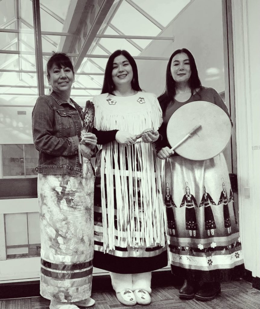 Three Indigenous women in ribbon skirts hold a feather, rattle, and drum, framed by sunlight streaming through a window.
