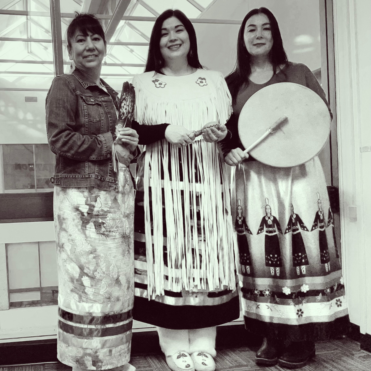 Three Indigenous women in ribbon skirts hold a feather, rattle, and drum, framed by sunlight streaming through a window.