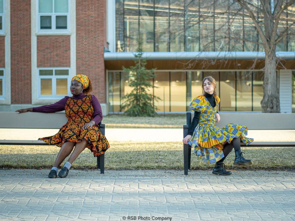 Two women, Non-African and African in vibrant African print dresses sit apart on benches, outdoors, in front of a building.