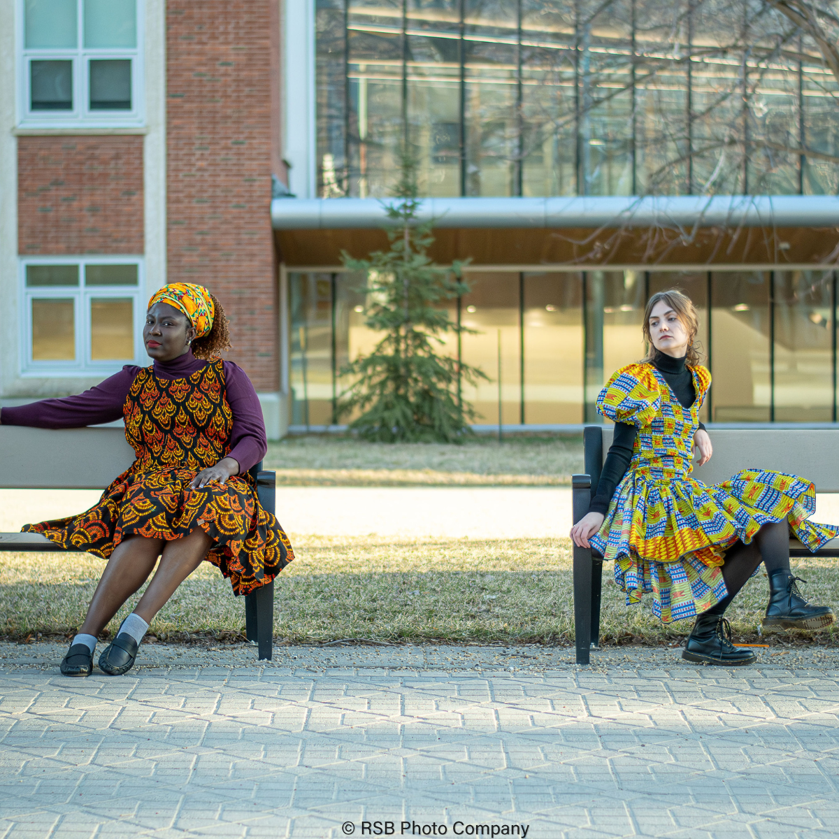 Two women, Non-African and African in vibrant African print dresses sit apart on benches, outdoors, in front of a building.