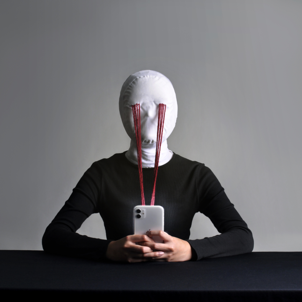 A person in a white mask sits at a table staring at a phone; red threads form direct lines from the mask’s eyes to the screen