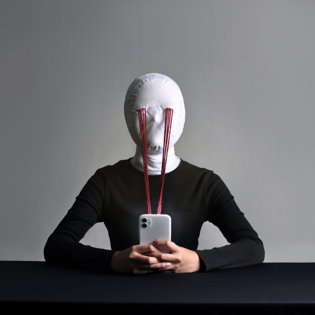 A person in a white mask sits at a table staring at a phone; red threads form direct lines from the mask’s eyes to the screen