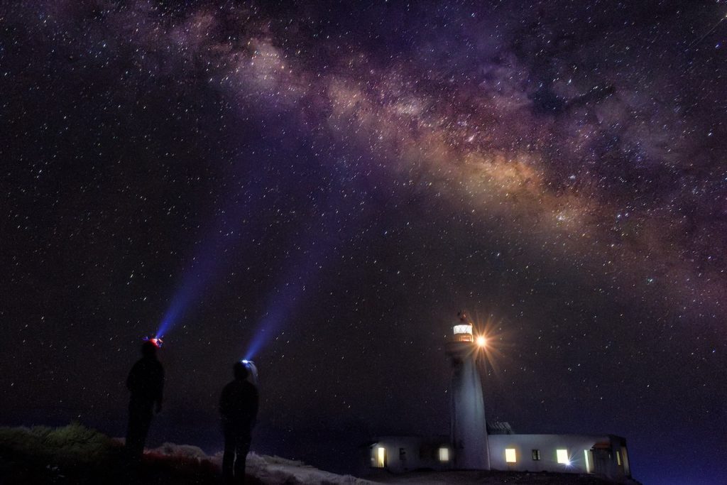 Two researchers stand before a lighthouse under a starry sky on a remote island, evoking solitude, connection, and climate ties.