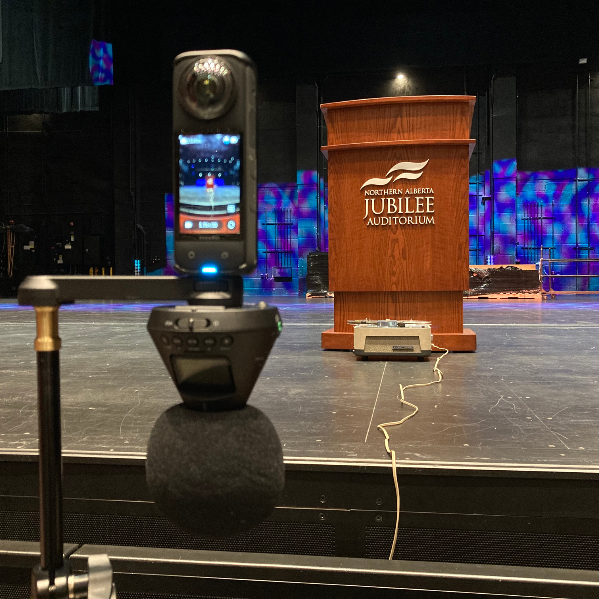 A 360 camera and a mic. In the background, a reel-to-reel tape player sits on a stage with a podium.