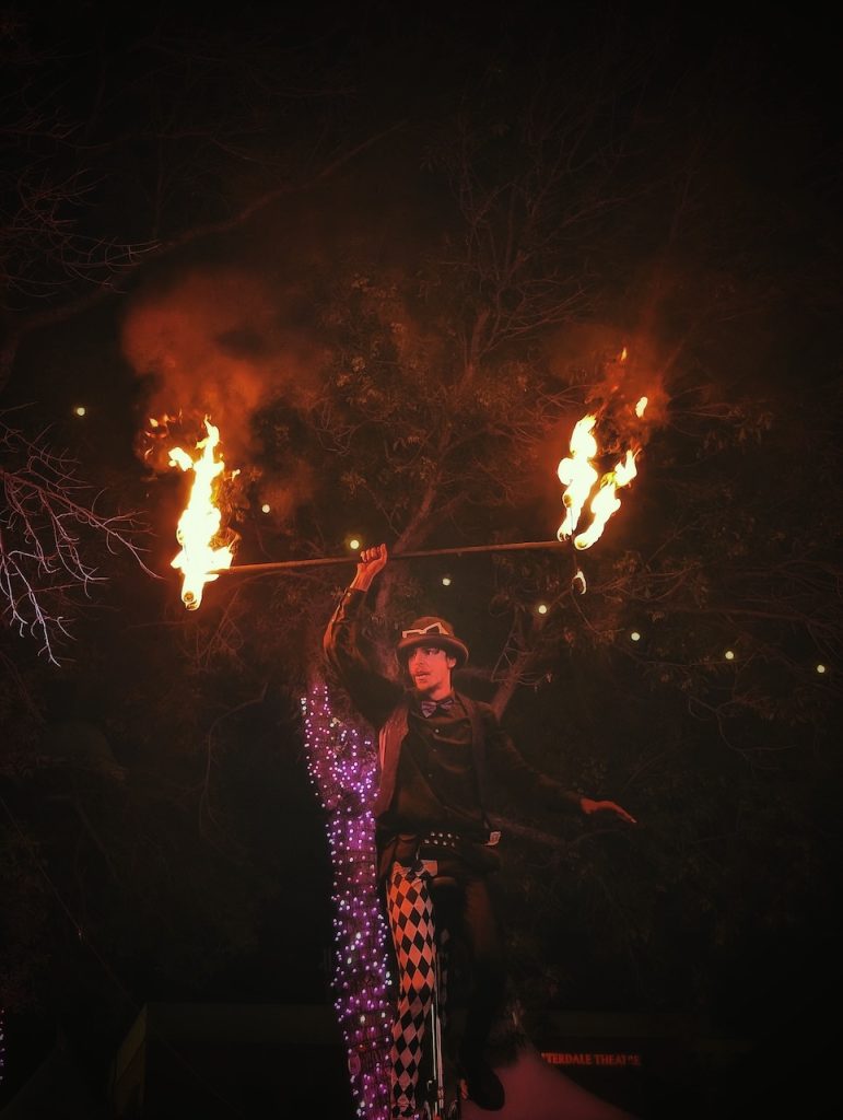 A street performer balances on a unicycle at night while holding a flaming staff.