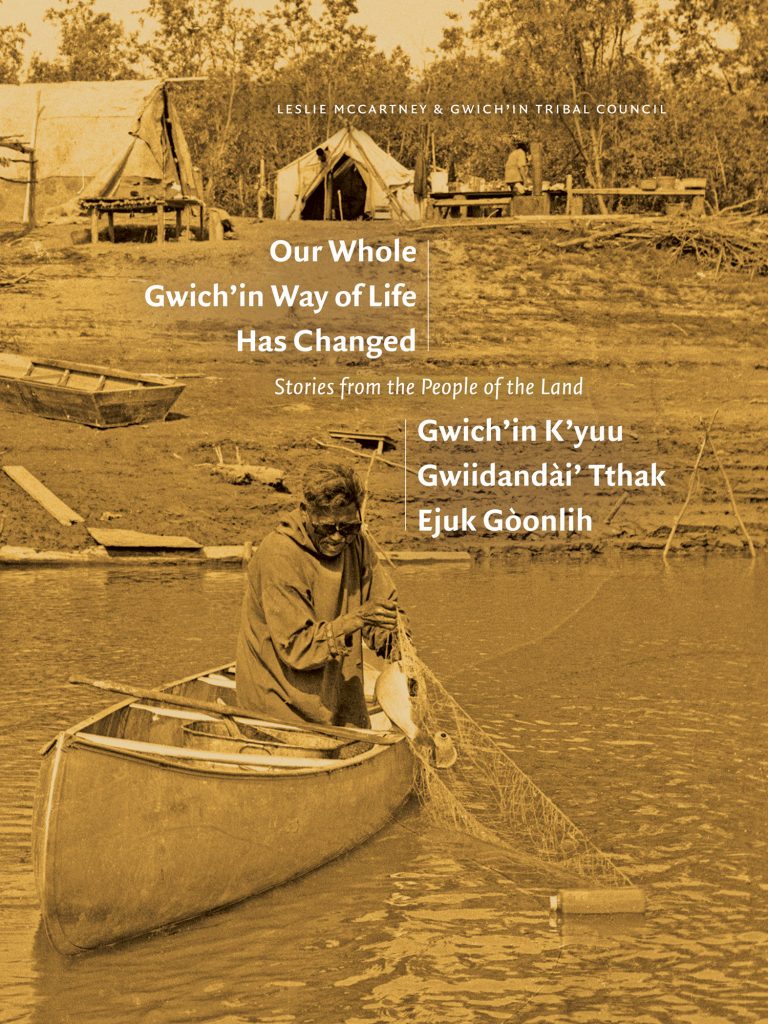 A sepia photograph by James Jerome from the NWT Archives featuring a fisherman in a canoe pulling in his fishing net with a medium-sized fish and a plastic bottle caught in the threads. The canoe is offshore, not far from the bank, and a fishing camp appears in the background with two visible tents and a large table where another person is standing. The fisherman is wearing sunglasses and a long-sleeved robe with patterns on the cuffs. The authors’ names feature at the top left corner in small white letters, and the title, in English and Gwichʼin language, features in medium-sized white letters at the centre of the cover just above the fisherman’s head.