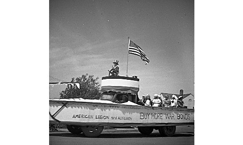 A parade float that looks like a boat, with an American flag. The float is carrying nurses, and reads 'American Legion and Auxiliary - Buy More War Bonds.'