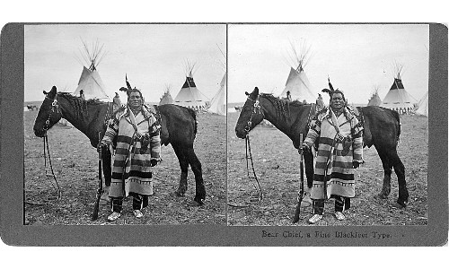 Bear Chief standing in front of a horse, rifle in hand, with tipis in the background. Text at the bottom of the stereoscope card reads 'Bear Chief, a fine Blackfeet Type.'