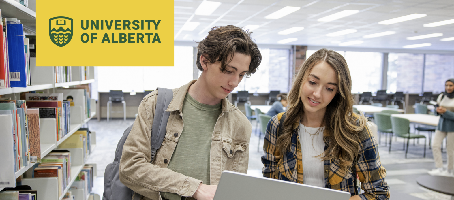 two students stand in a well lit library looking happily at a laptop